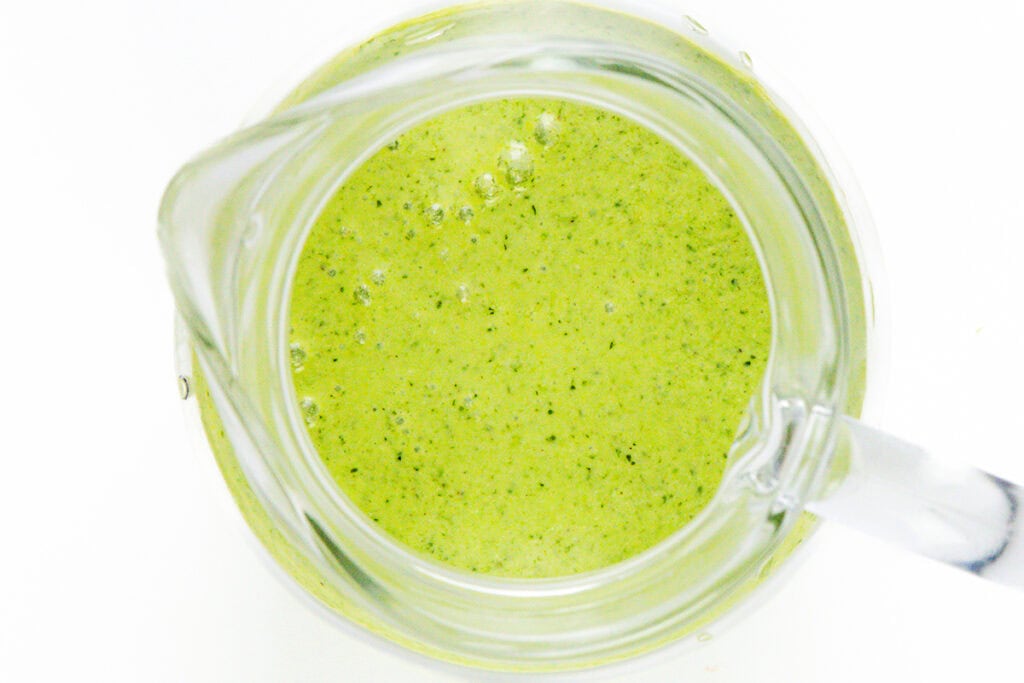 Top view of a glass pitcher filled with frothy green cilantro dressing, possibly for a couscous salad, set against a white background.