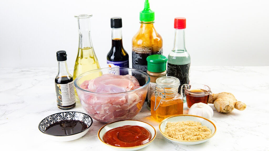 A variety of ingredients for cooking Sticky Asian BBQ Chicken Thighs are arranged on a white surface, including a bowl of raw chicken, bottles of sauces, fresh ginger, garlic, small bowls with ketchup, soy sauce, brown sugar, and a jar of honey.