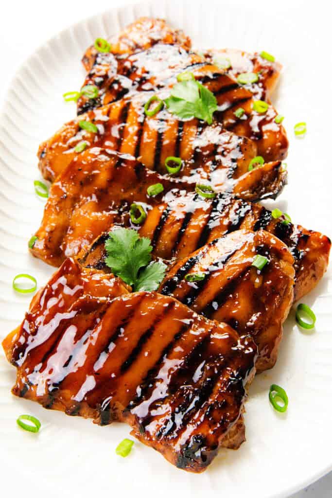 Grilled chicken breasts glazed with a shiny teriyaki sauce, inspired by Sticky Asian BBQ Chicken Thighs, garnished with chopped green onions and fresh cilantro, served on a white plate.