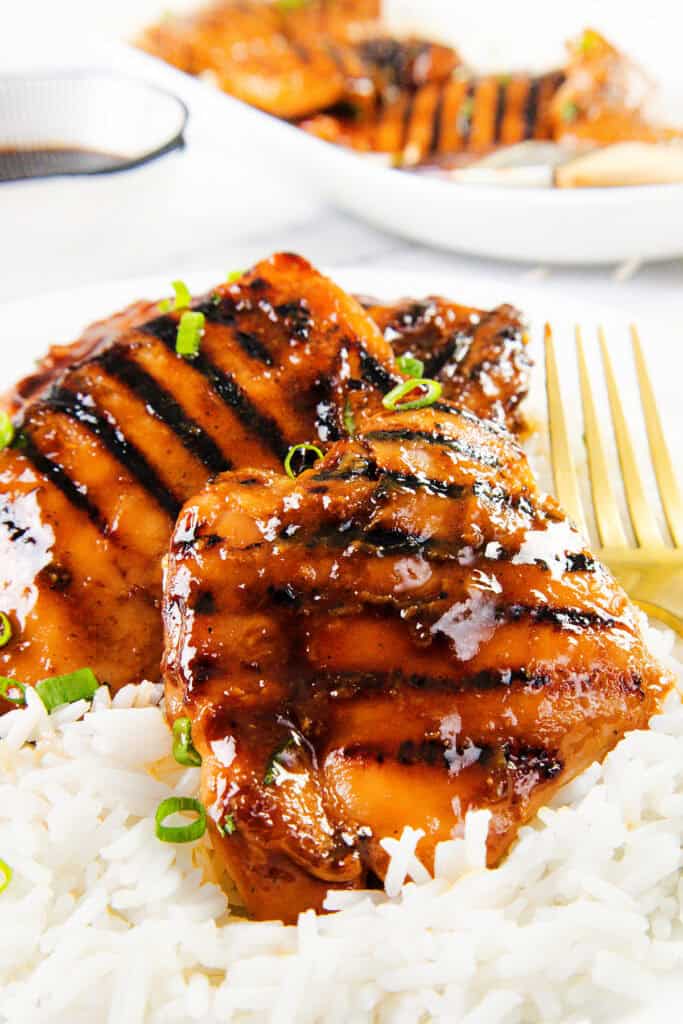 Grilled glazed chicken breasts with grill marks served on a bed of white rice, garnished with chopped green onions. A gold fork rests beside the chicken, evoking flavors reminiscent of Sticky Asian BBQ Chicken Thighs. More grilled chicken in the background.