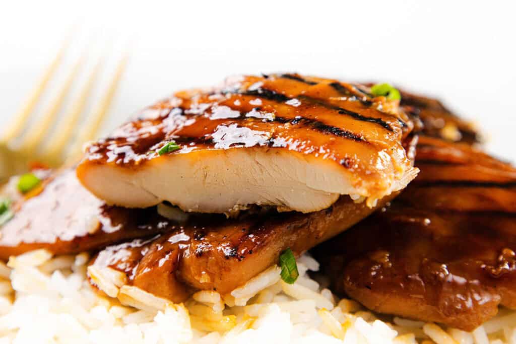 Close-up of grilled Asian BBQ Chicken Thighs slices with a glossy sauce, served on a bed of white rice. A fork is visible in the background. The chicken has visible grill marks and is garnished with herbs.