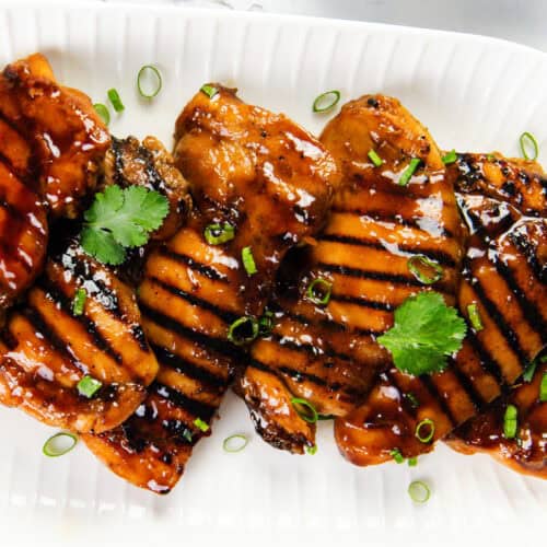 Grilled Asian BBQ Chicken breasts glazed with a shiny sauce, garnished with fresh cilantro leaves and chopped green onions, arranged on a white ridged platter.