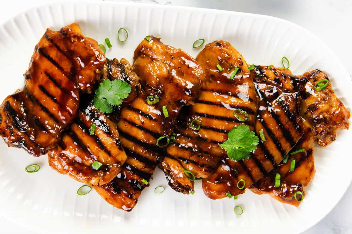 Grilled Asian BBQ Chicken breasts glazed with a shiny sauce, garnished with fresh cilantro leaves and chopped green onions, arranged on a white ridged platter.