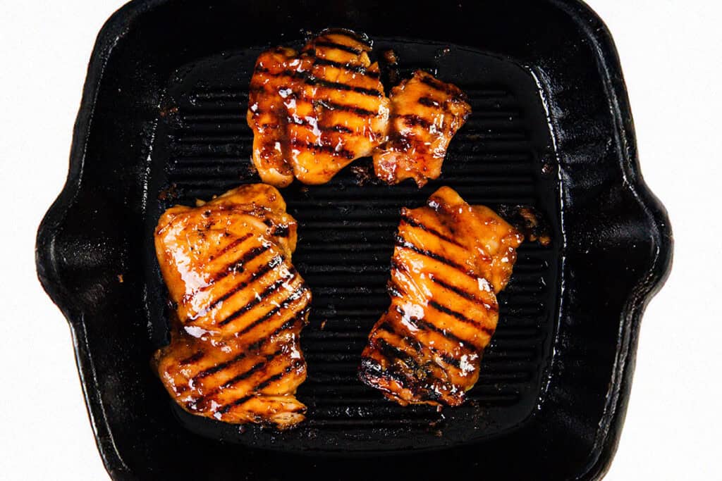 Three pieces of Sticky Asian BBQ Chicken Thighs with dark grill marks are cooking on a black cast iron grill pan, viewed from above against a white background.