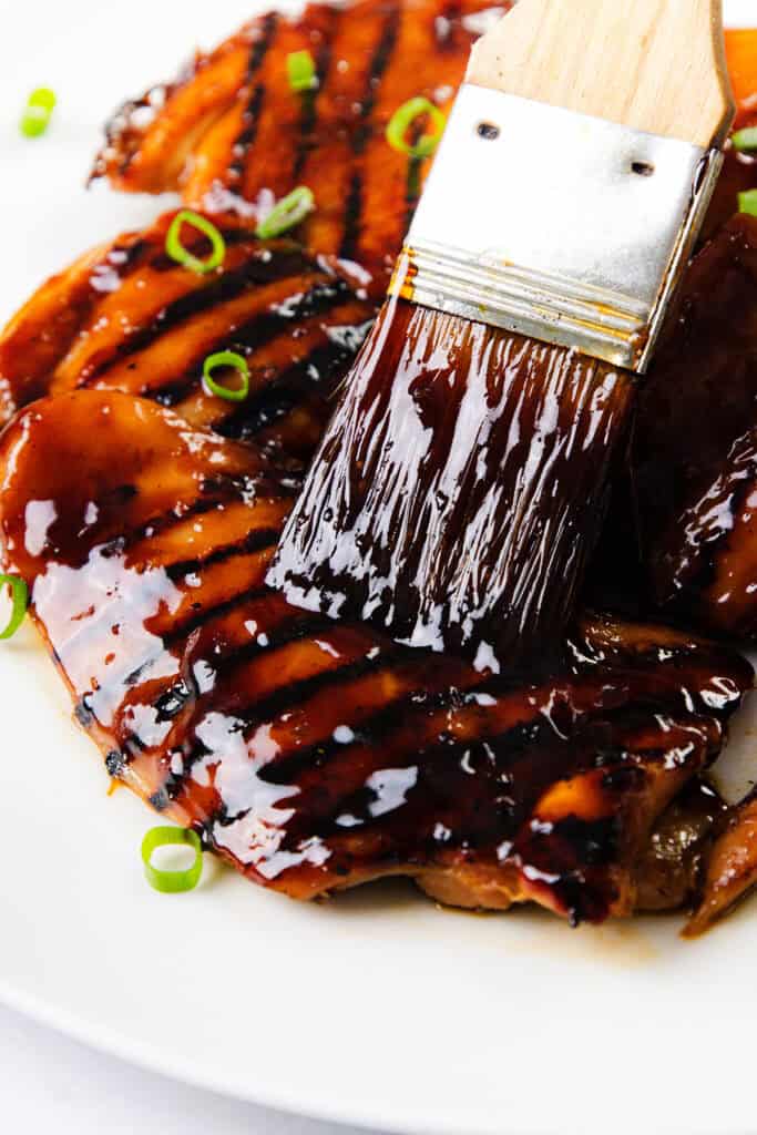 A close-up of a basting brush coating Sticky Asian BBQ Chicken Thighs with a glossy barbecue sauce, garnished with sliced green onions on a white plate.