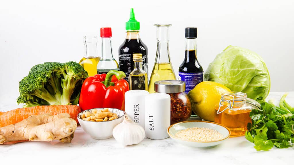 A variety of fresh vegetables, oils, sauces, and spices are arranged on a white surface—perfect ingredients for an Asian Roasted Broccoli Salad or a homemade Sesame Vinaigrette.