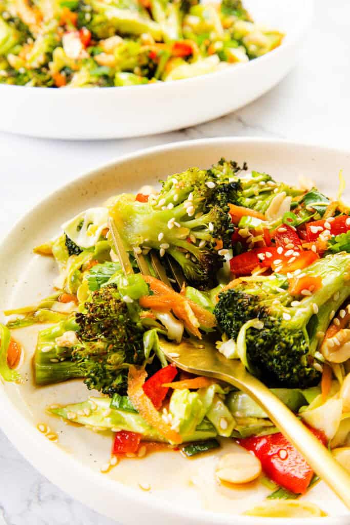 A plate of vibrant stir-fried vegetables, including broccoli, red bell pepper, and shredded carrots, garnished with sesame seeds and peanuts—reminiscent of an Asian Roasted Broccoli Salad—with a fork resting on the plate.