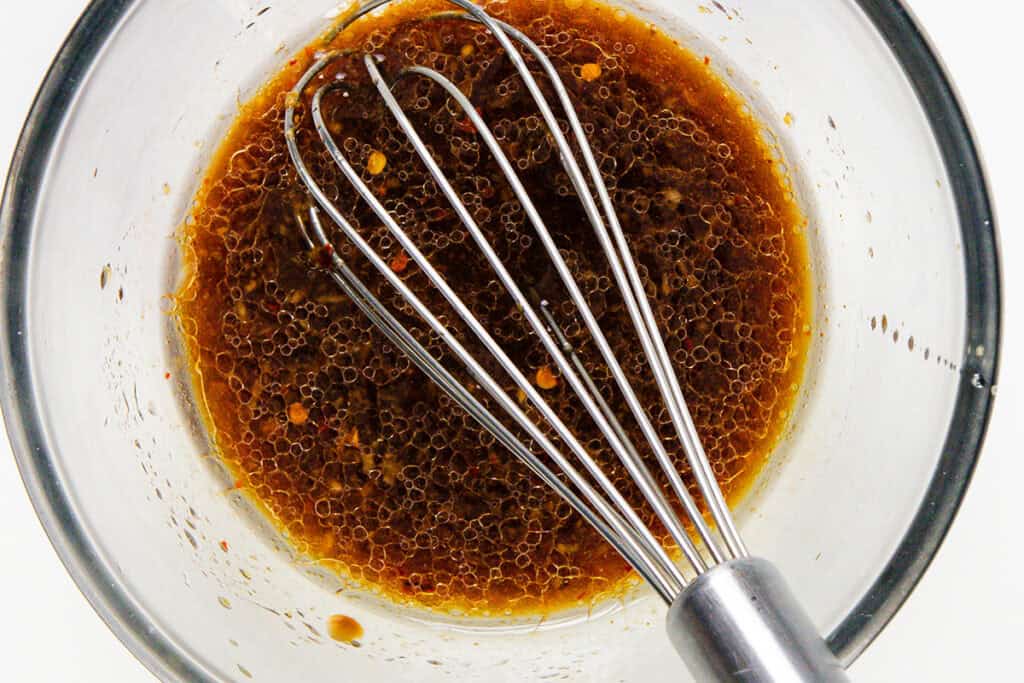 A metal whisk rests in a glass bowl containing a brown, oily, and slightly bubbly Sesame Vinaigrette mixture, viewed from above on a white background.