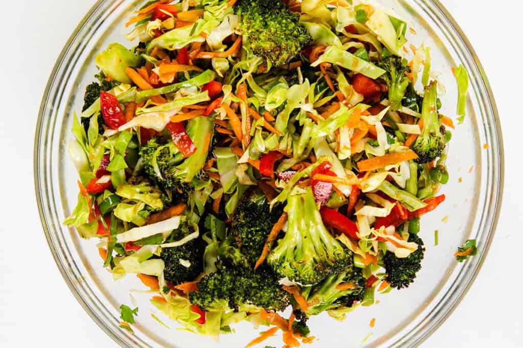 A glass bowl filled with a colorful Asian Roasted Broccoli Salad featuring shredded carrots, red bell peppers, and chopped cabbage, all tossed in a savory sesame vinaigrette on a white background.