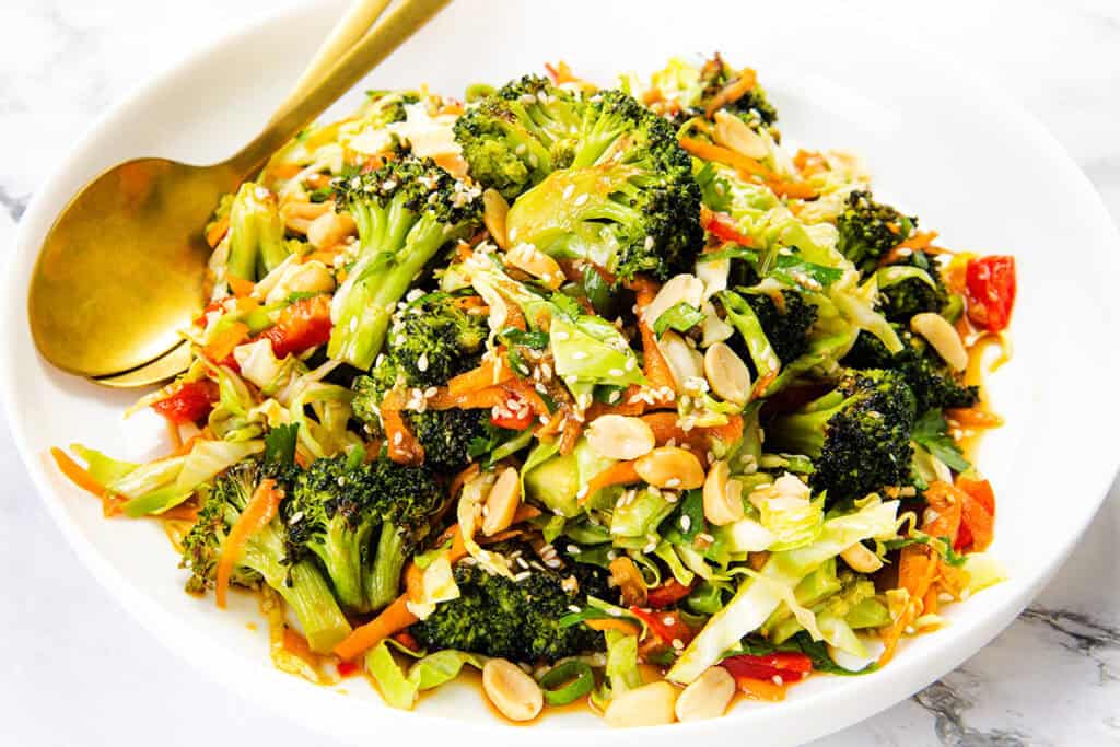 A white bowl filled with a colorful Asian Roasted Broccoli Salad—broccoli, shredded carrots, cabbage, red bell peppers, peanuts, and sesame seeds—tossed with sesame vinaigrette. A gold serving spoon rests on the side of the bowl.