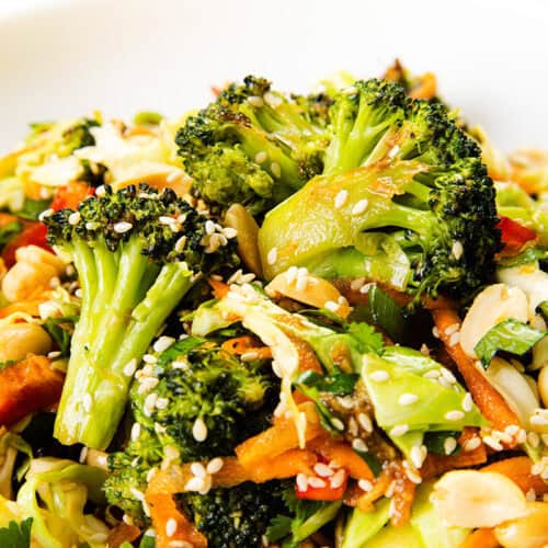 A close-up of a vibrant Asian Roasted Broccoli Salad with shredded carrots, red bell peppers, sliced almonds, and sesame seeds, all tossed in a flavorful sesame vinaigrette on a white plate.