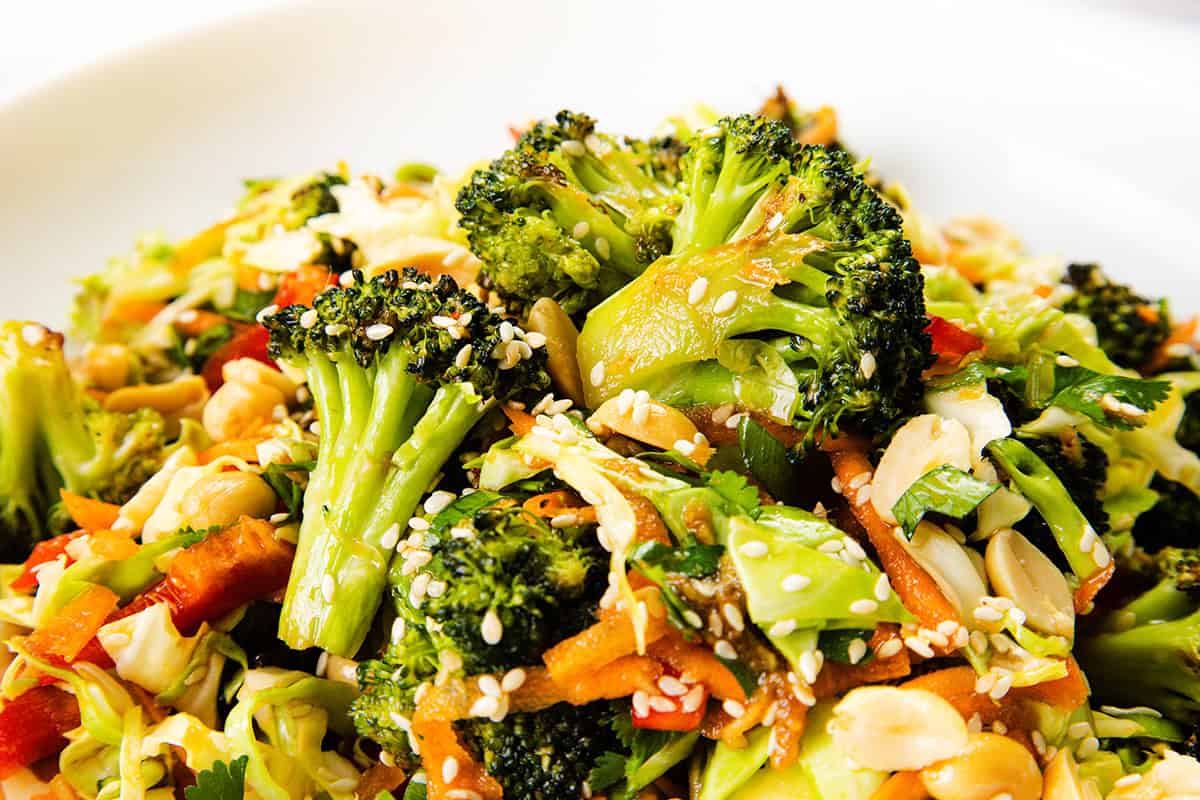 A close-up of a vibrant Asian Roasted Broccoli Salad with shredded carrots, red bell peppers, sliced almonds, and sesame seeds, all tossed in a flavorful sesame vinaigrette on a white plate.