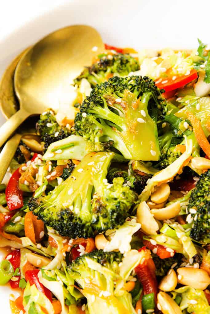 A close-up of an Asian Roasted Broccoli Salad featuring roasted broccoli, sliced red peppers, carrots, green onions, peanuts, and sesame seeds in a white bowl with gold utensils, finished with a tangy sesame vinaigrette.