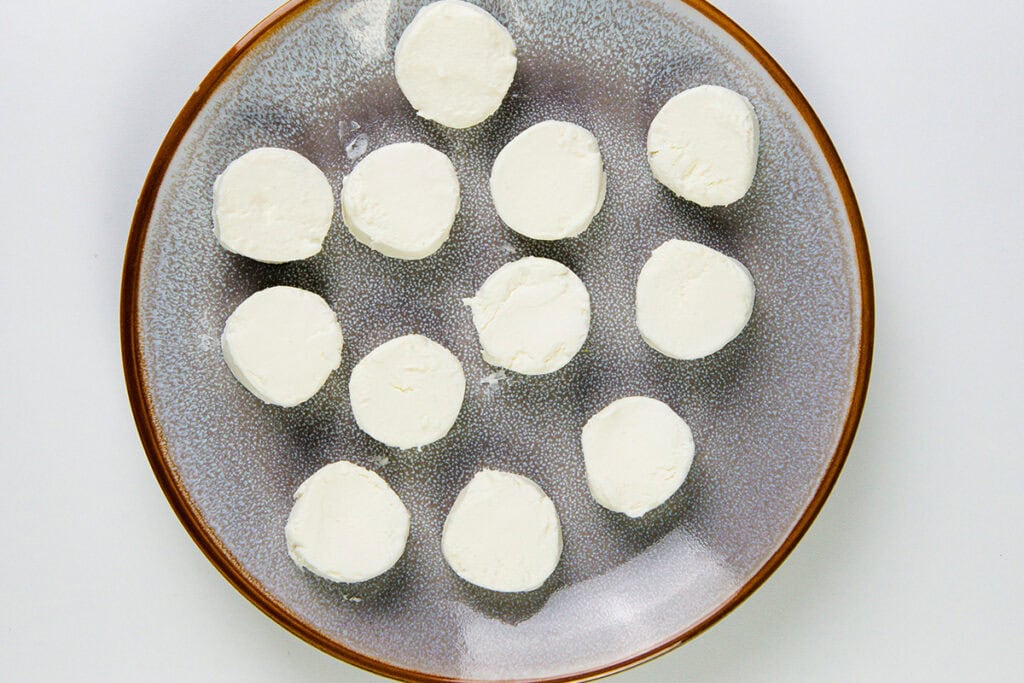 A brown-rimmed, gray plate with thirteen round slices of fried goat cheese arranged in a loose pattern on a white background.