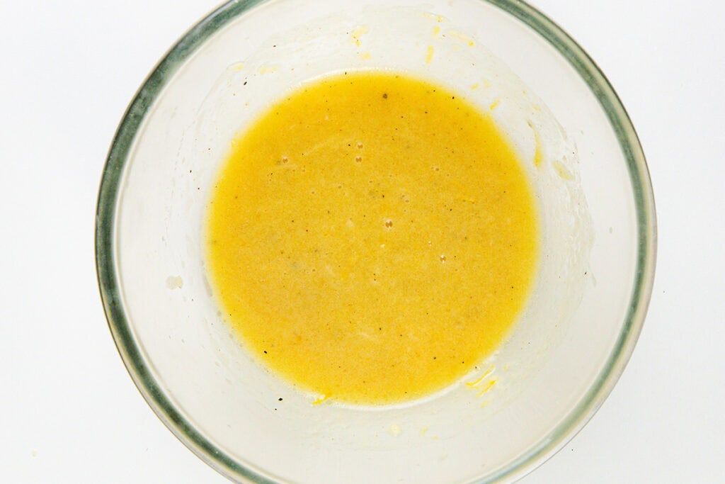 A clear glass bowl containing a yellow liquid mixture with a slightly grainy texture, likely infused with honey and herbs, viewed from above, set against a white background.