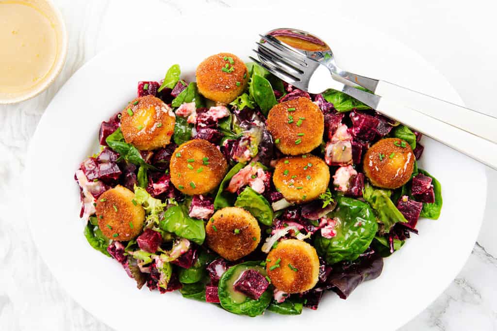 A white plate holds a vibrant salad of leafy greens, diced beets, and feta cheese, highlighted by crispy fried goat cheese rounds. A fork and spoon rest nearby, with a small bowl of dressing on the side.