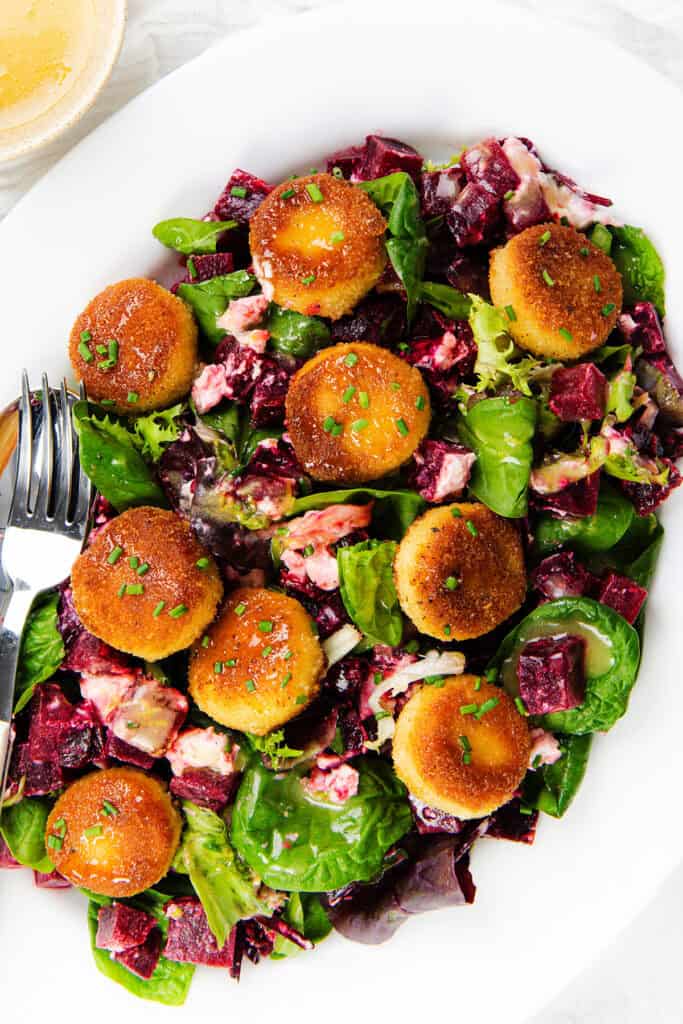 A white plate with a fresh Beet Goat Cheese Salad—mixed greens, diced beets, creamy herb dressing, and eight golden-brown breaded goat cheese rounds drizzled with honey, with two forks resting on the side.
