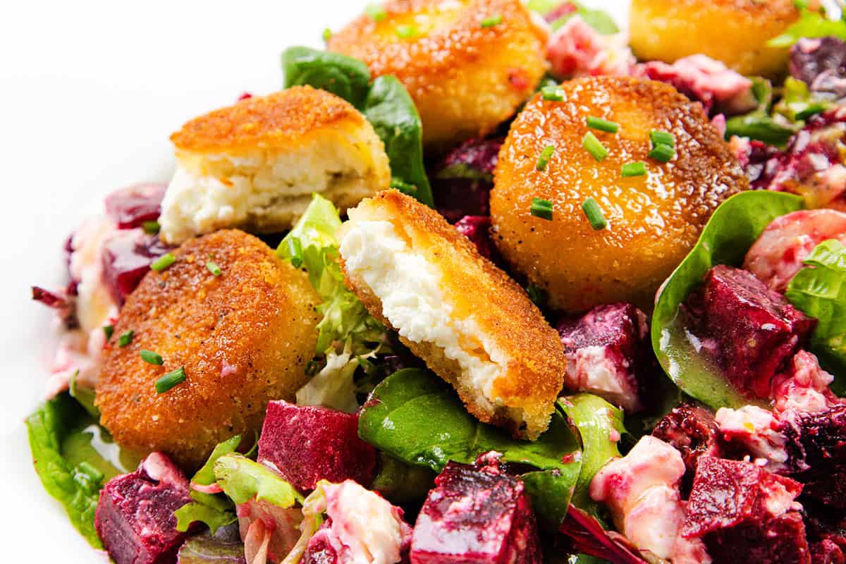 A close-up of a salad with crispy Fried Goat Cheese medallions, mixed greens, and cubed roasted beets, all garnished with chopped chives.