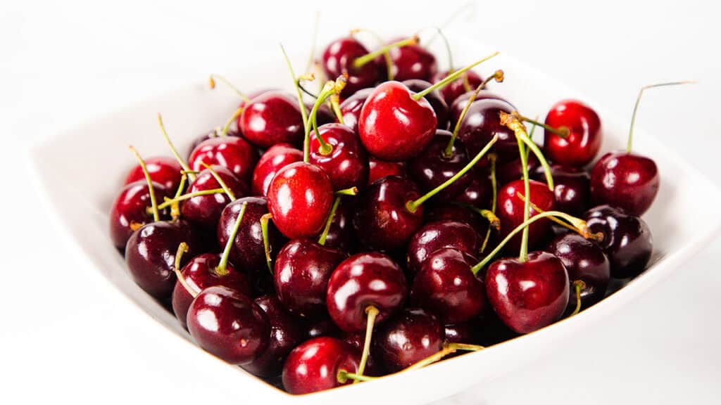 A white square bowl filled with fresh, ripe red cherries with green stems, perfect for creating a delectable Flaming Cherries Jubilee dessert, is placed on a white surface.