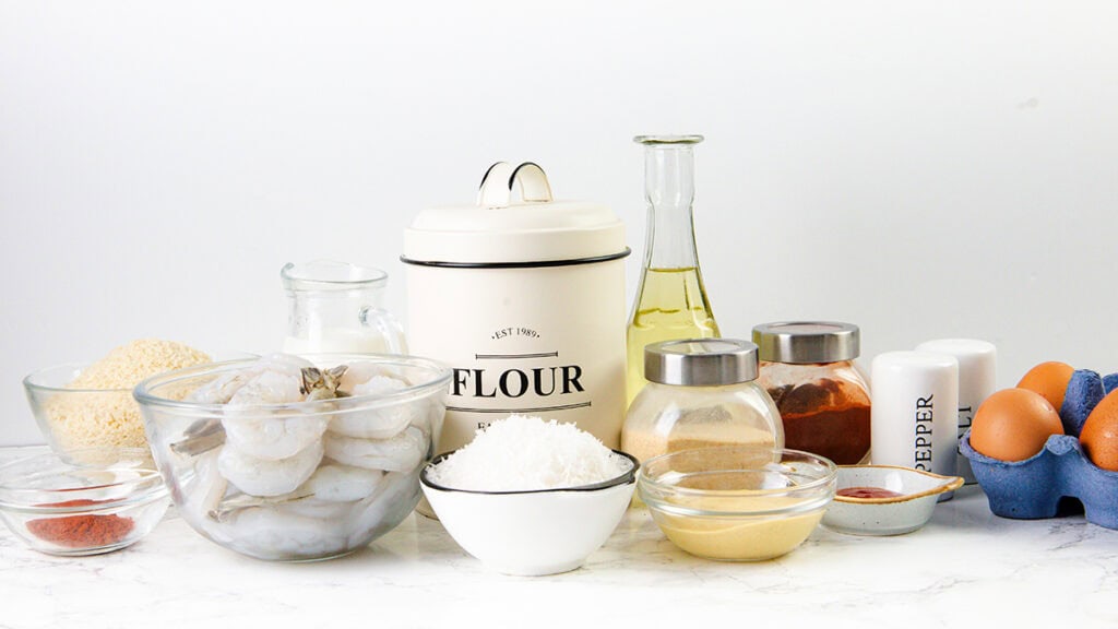 Various cooking ingredients are arranged on a white surface, including raw shrimp in a bowl, flour, eggs, spices in jars, milk, oil, grated cheese, a salt shaker, and a pepper shaker—perfect for making Coconut Shrimp or your favorite Shrimp Recipe.