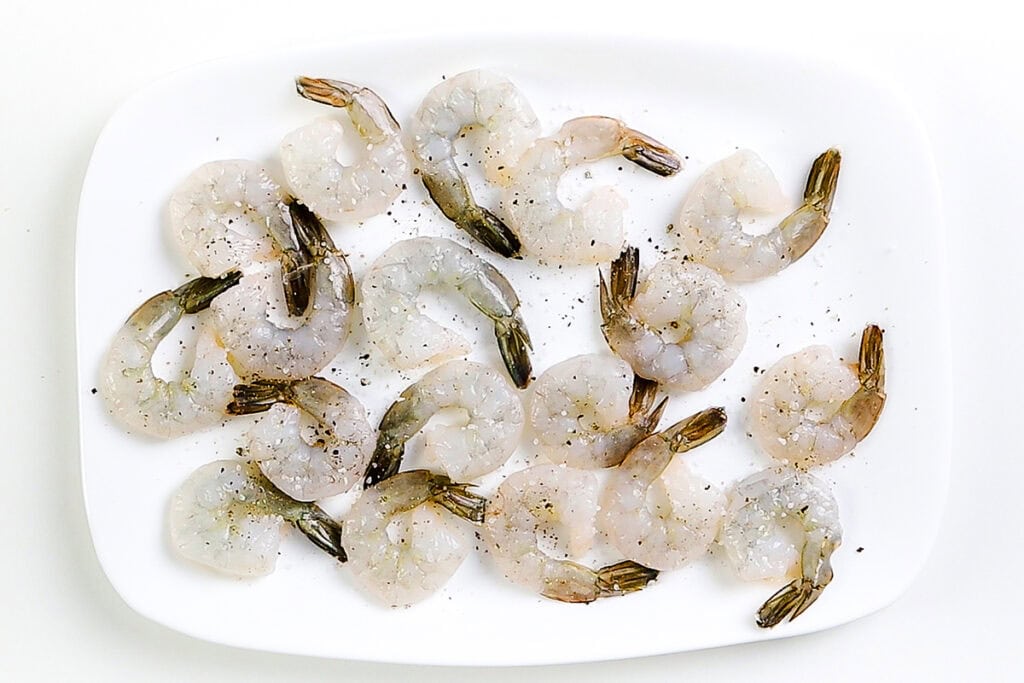 Raw shrimp with tails on, sprinkled with black pepper, arranged on a white plate against a white background—perfect for your next coconut shrimp or sweet chili dip shrimp recipe.