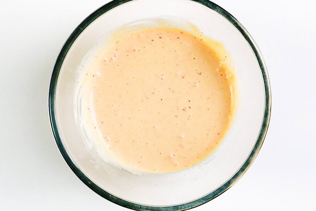 A glass bowl filled with a creamy, pale orange Sweet Chili Dip speckled with small herbs and spices, placed on a white surface—perfect for pairing with Coconut Shrimp.
