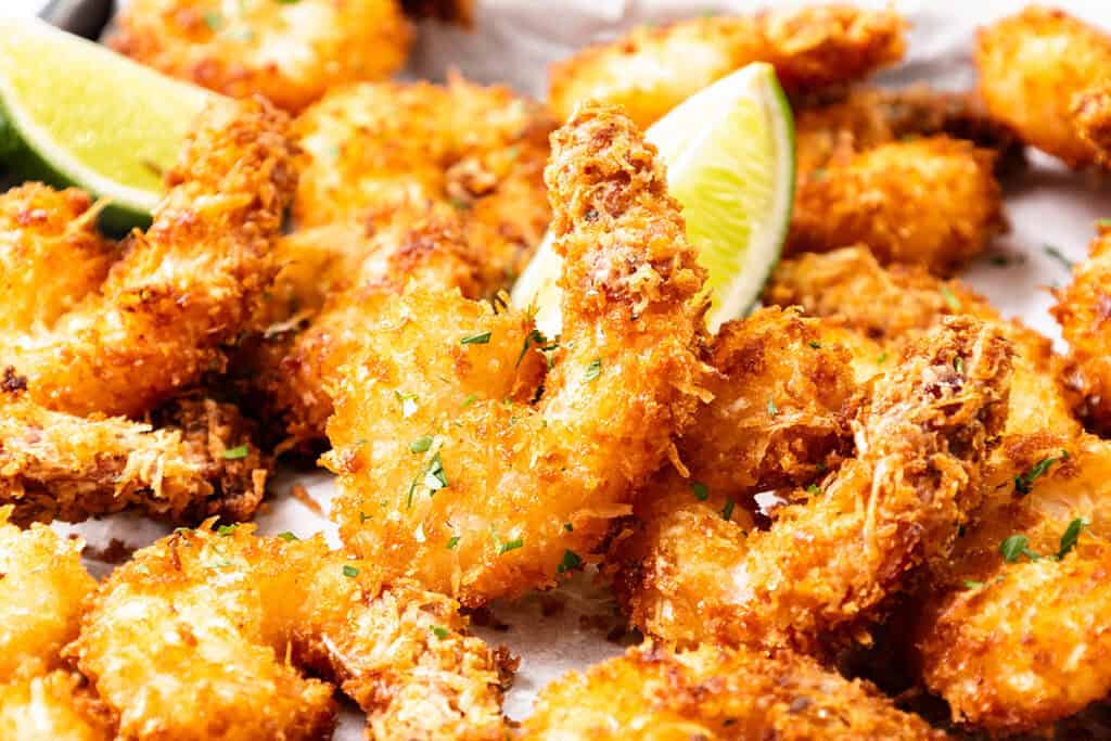 Golden, crispy Coconut Shrimp garnished with chopped herbs and served with lime wedges on a white surface—perfect for dipping in sweet chili dip.
