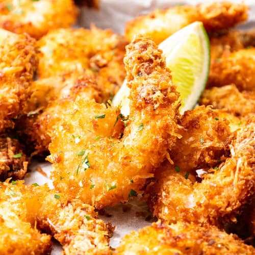 Golden, crispy Coconut Shrimp garnished with chopped herbs and served with lime wedges on a white surface—perfect for dipping in sweet chili dip.