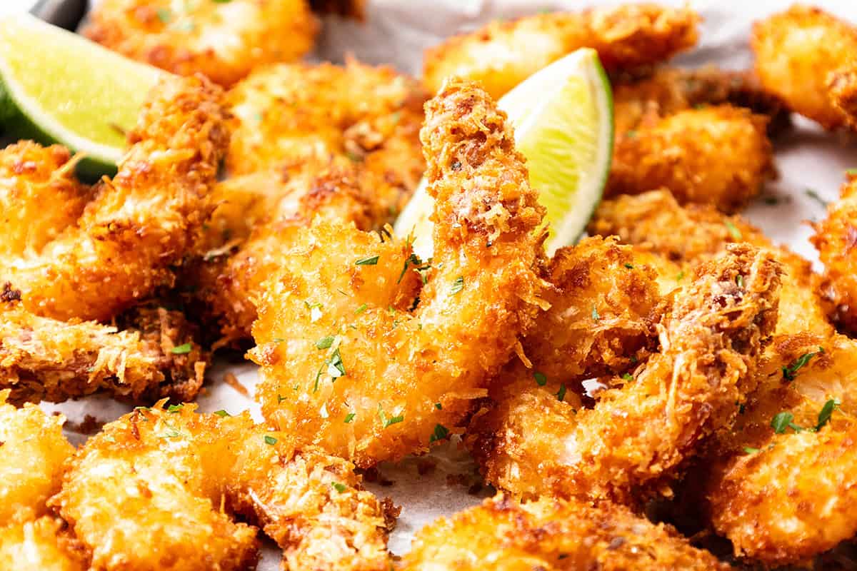 Golden, crispy Coconut Shrimp garnished with chopped herbs and served with lime wedges on a white surface—perfect for dipping in sweet chili dip.