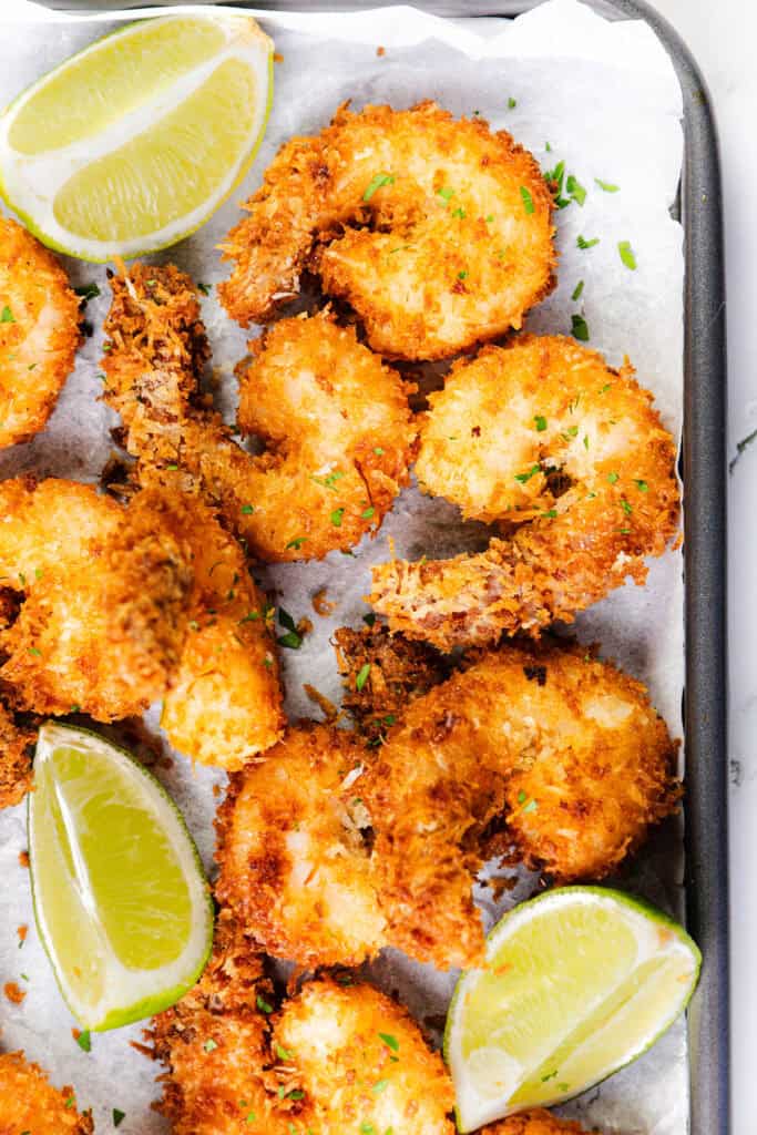 Golden, crispy coconut shrimp garnished with chopped herbs, served on parchment paper with fresh lime wedges and a side of sweet chili dip.