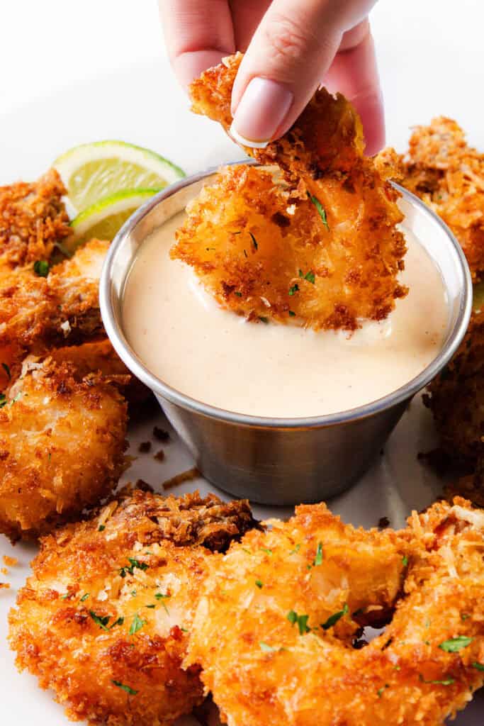 A hand dips crispy coconut shrimp into a cup of sweet chili dip, with more shrimp and lime wedges arranged on a white plate.