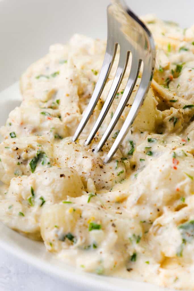 A close-up of a fork poised over creamy potato salad mixed with chopped herbs, cracked black pepper, and a rich, white dressing—evoking the decadent appeal of creamy gnocchi.