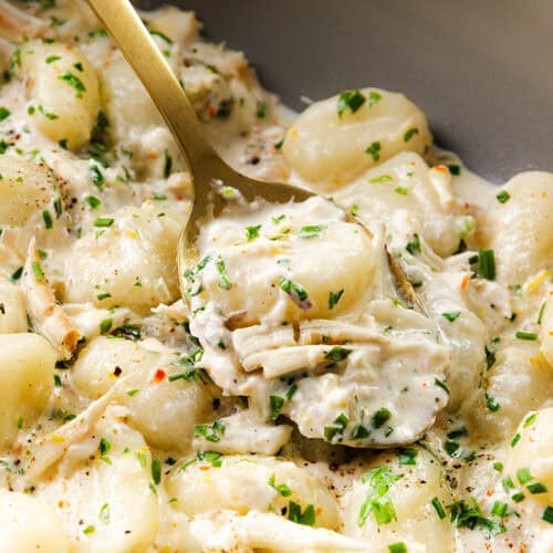 A close-up of creamy crab gnocchi with shredded chicken, garnished with chopped herbs, served with a gold spoon scooping up a portion.
