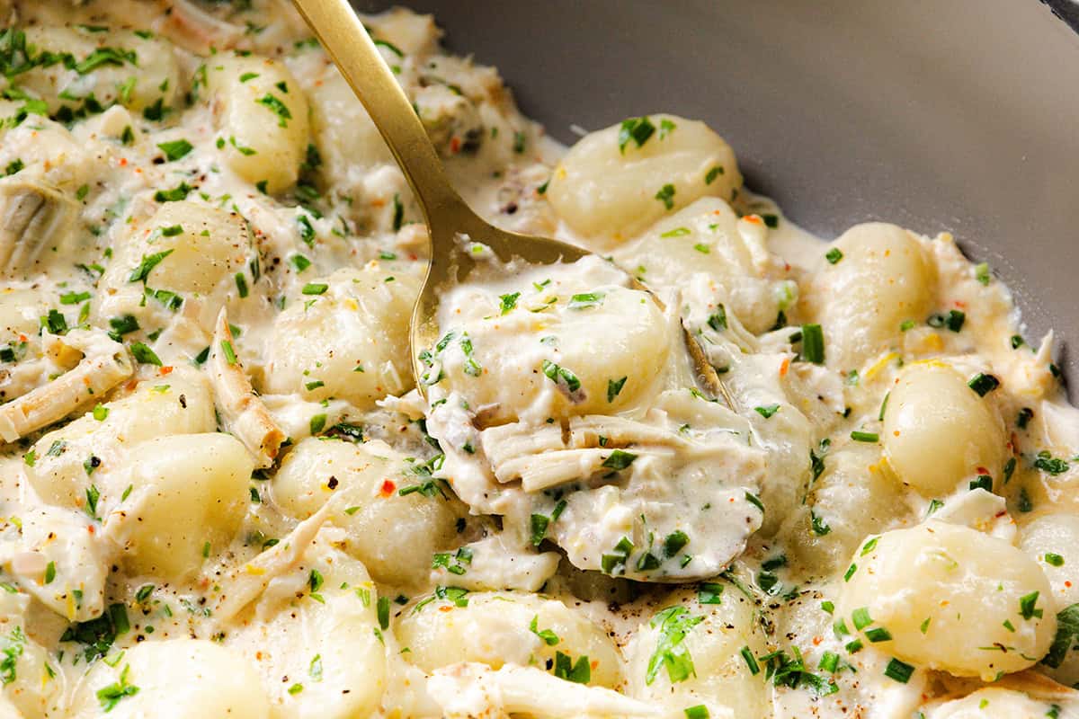 A close-up of creamy crab gnocchi with shredded chicken, garnished with chopped herbs, served with a gold spoon scooping up a portion.