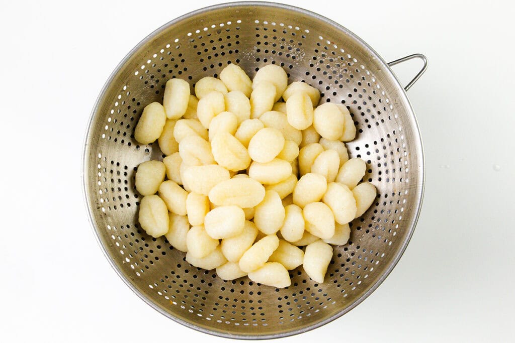 A metal colander filled with creamy gnocchi sits on a white surface. The gnocchi are small, oval-shaped, and pale, indicating they are freshly prepared.