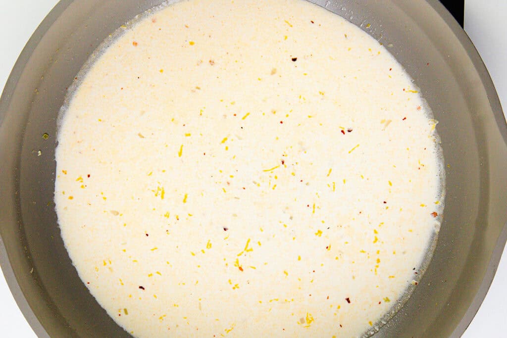 A close-up view of a creamy white sauce for crab gnocchi, with small yellow zest pieces and specks of seasoning in a gray pan.