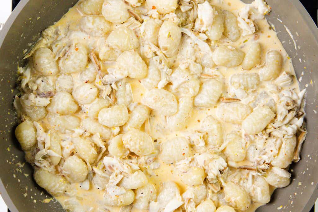 Creamy gnocchi mixed with shredded chicken in a light-colored sauce, cooked in a large skillet. This crab gnocchi variation offers a rich, hearty texture with visible bits of chicken throughout.