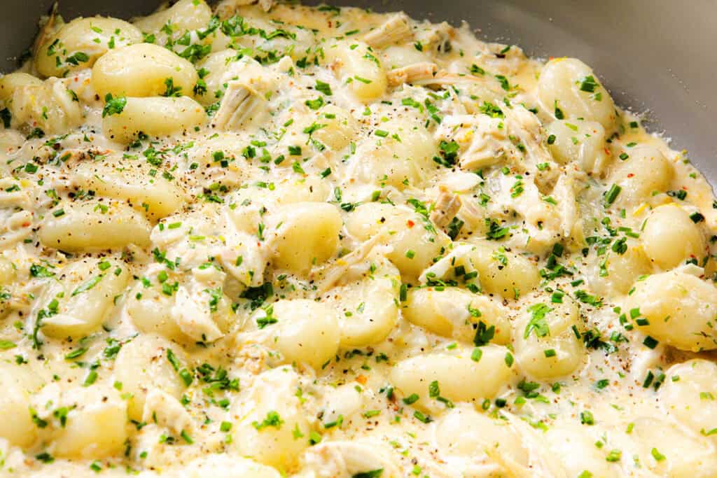 Creamy gnocchi dish with shredded chicken, garnished with chopped chives and cracked black pepper, served in a skillet. The rich sauce evenly coats the gnocchi and chicken, reminiscent of a decadent crab gnocchi or seafood pasta experience.