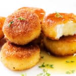 Five golden-brown, breaded fried goat cheese rounds are stacked on a white plate, with one cut open to reveal the creamy inside. They are garnished with fresh herbs and drizzled with honey.