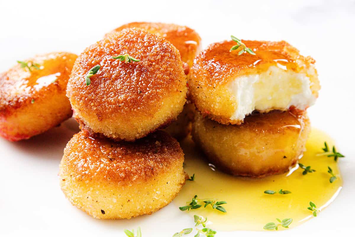 Five golden-brown, breaded fried goat cheese rounds are stacked on a white plate, with one cut open to reveal the creamy inside. They are garnished with fresh herbs and drizzled with honey.