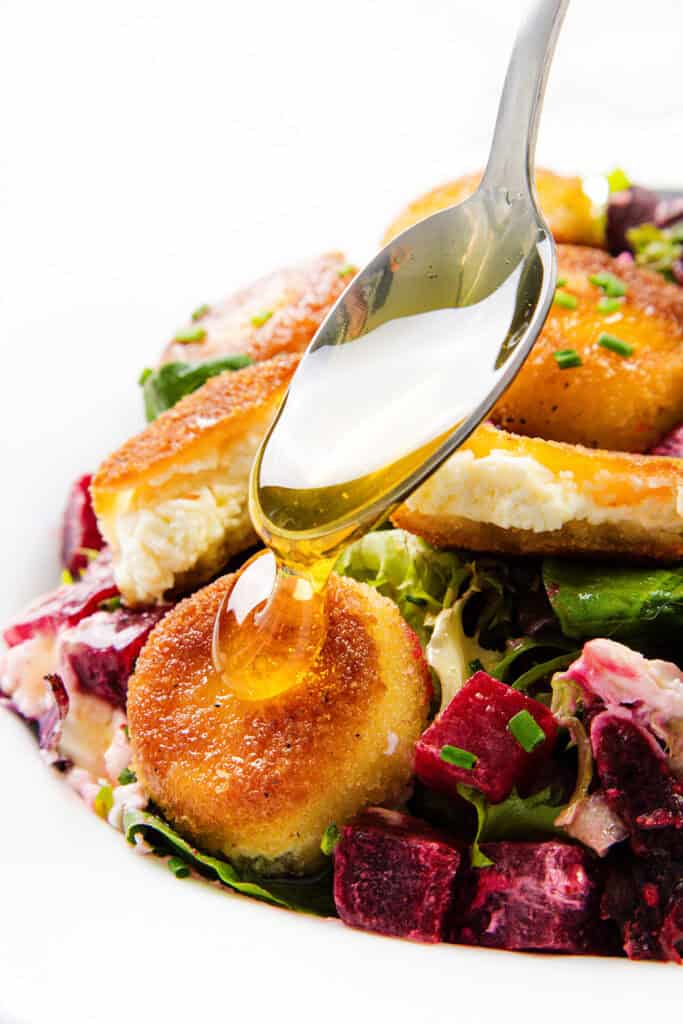 A close-up of a spoon drizzling honey over crispy fried goat cheese rounds on a salad with mixed greens and diced beets.