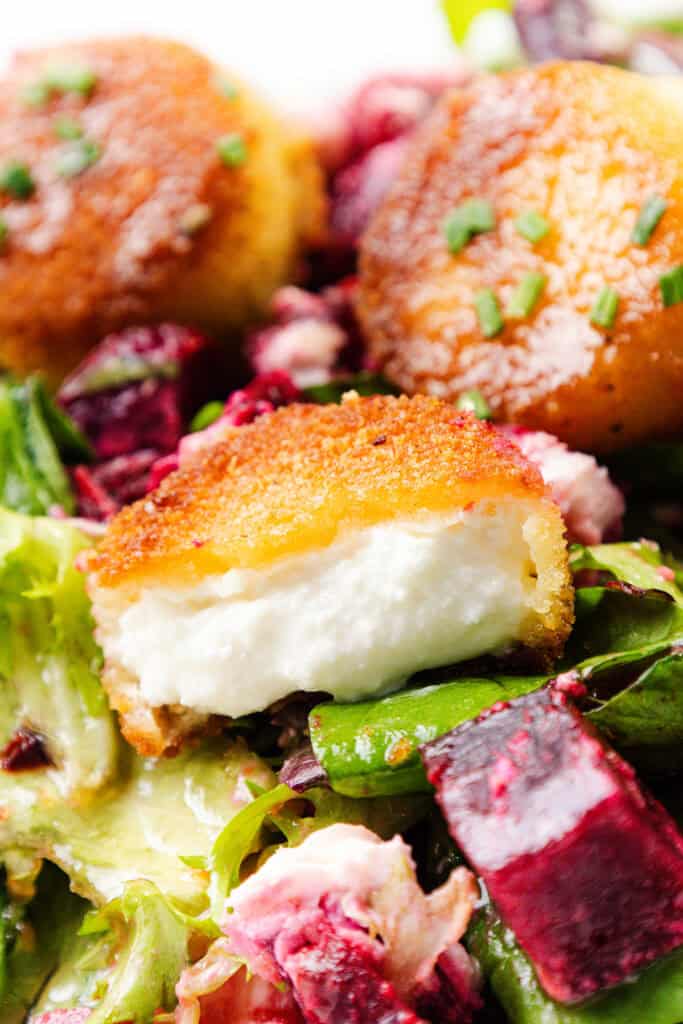 Close-up of a salad with mixed greens, roasted beet cubes, and crispy fried goat cheese rounds, one of which is cut open to reveal the creamy interior.