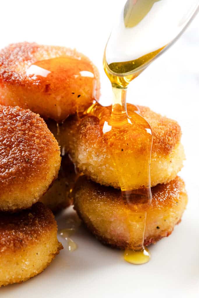 Golden syrup is being poured over a stack of round, crispy Fried Goat Cheese fritters on a white surface, creating a glossy, appetizing finish.