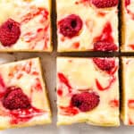 Six Raspberry Cheesecake Blondies, cut into squares, shown close-up. Each bar has a creamy white cheesecake layer with swirls of raspberry sauce and whole raspberries on top, resting on white parchment paper.