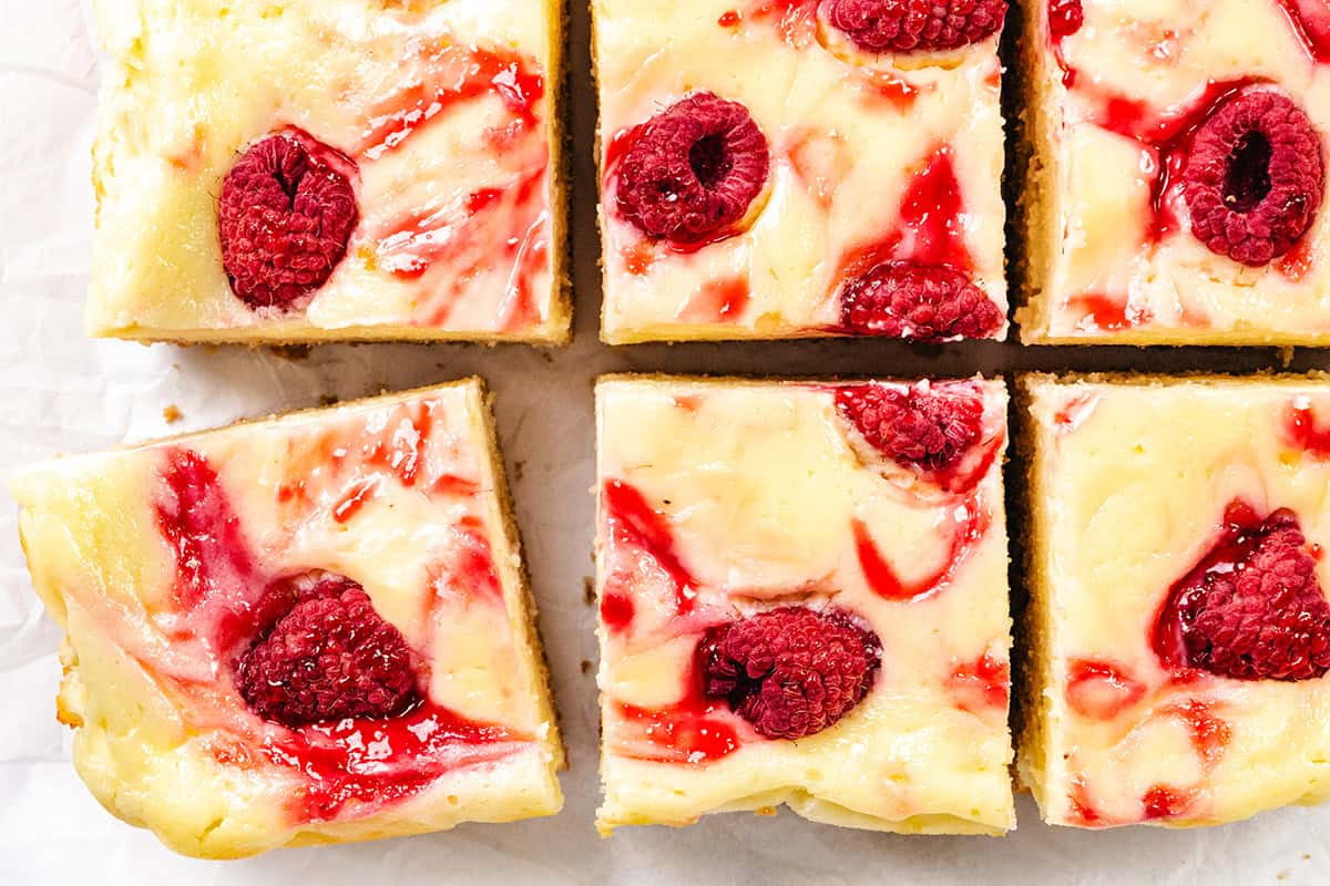Six Raspberry Cheesecake Blondies, cut into squares, shown close-up. Each bar has a creamy white cheesecake layer with swirls of raspberry sauce and whole raspberries on top, resting on white parchment paper.