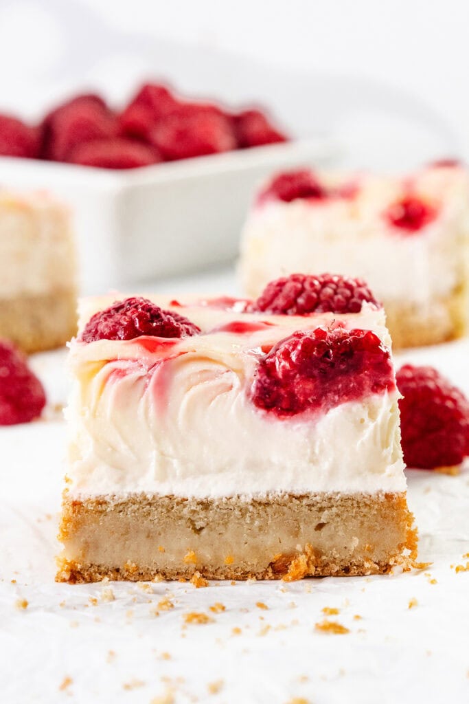 A close-up of Raspberry Cheesecake Blondies with a golden crust, creamy filling, and swirls of raspberry sauce topped with whole raspberries. More cheesecake bars and a dish of raspberries are blurred in the background.