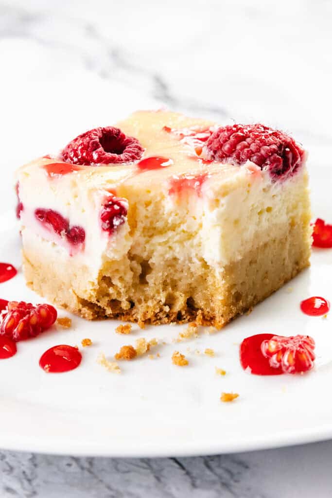 A square slice of raspberry cheesecake bar with a bite taken out, topped with raspberries and red sauce, sits on a white plate alongside buttery blondie crumbs and a drizzle of raspberry sauce.