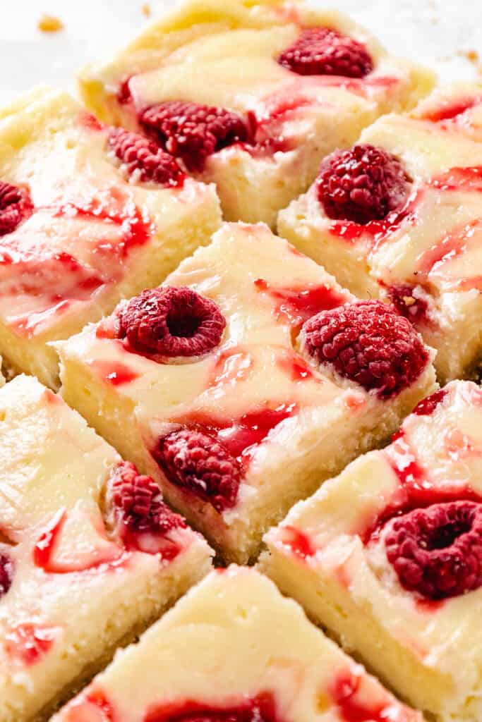 Close-up of Raspberry Cheesecake Blondies, cut into squares, topped with swirls of raspberry sauce and fresh raspberries. The creamy bars are arranged closely together on a white surface.