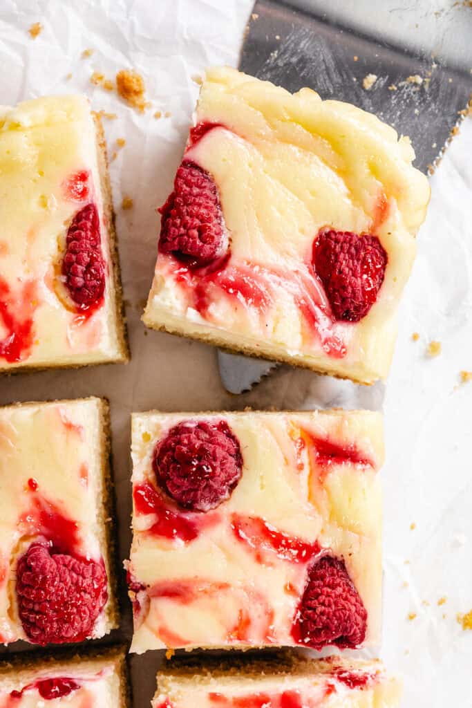 Top view of Raspberry Cheesecake Blondies swirled with raspberry sauce and topped with fresh raspberries, cut into squares on parchment paper, with a metal spatula lifting one piece.