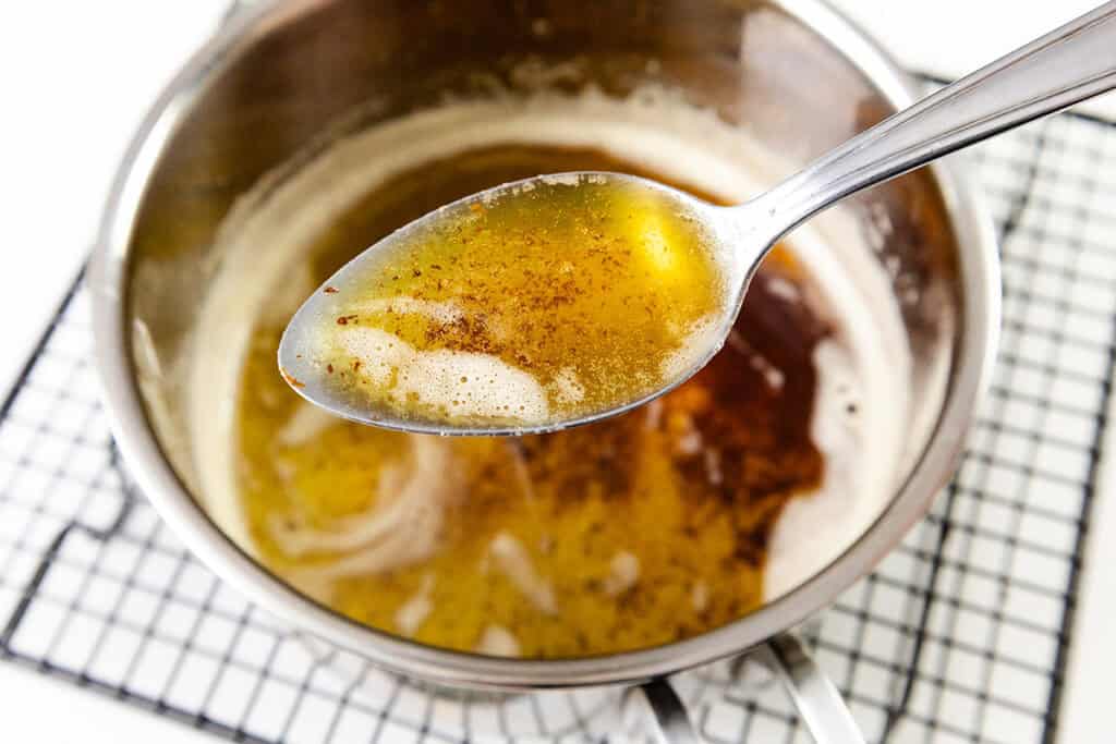 A close-up of a spoon holding browned butter with foamy bits, above a saucepan filled with more browned butter—perfect for adding rich flavor to cheesecake or blondies—resting on a black wire cooling rack.